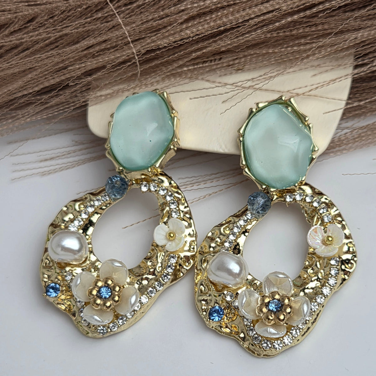 A pair of gold-toned statement earrings with large light green oval stones at the top, dangling scalloped gold chains, faux pearl beads, and small blue rhinestone accents, displayed on a white surface with brown hair in the background.